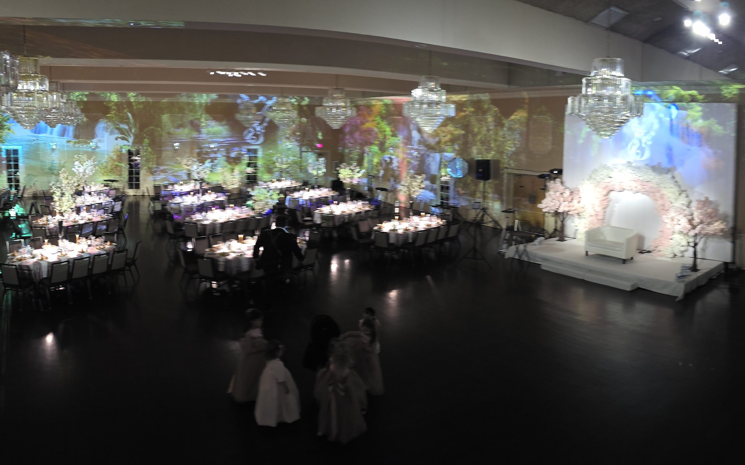The whole room from the balcony: the garden projected on every wall, the tables set, the blossom chuppah at the far end