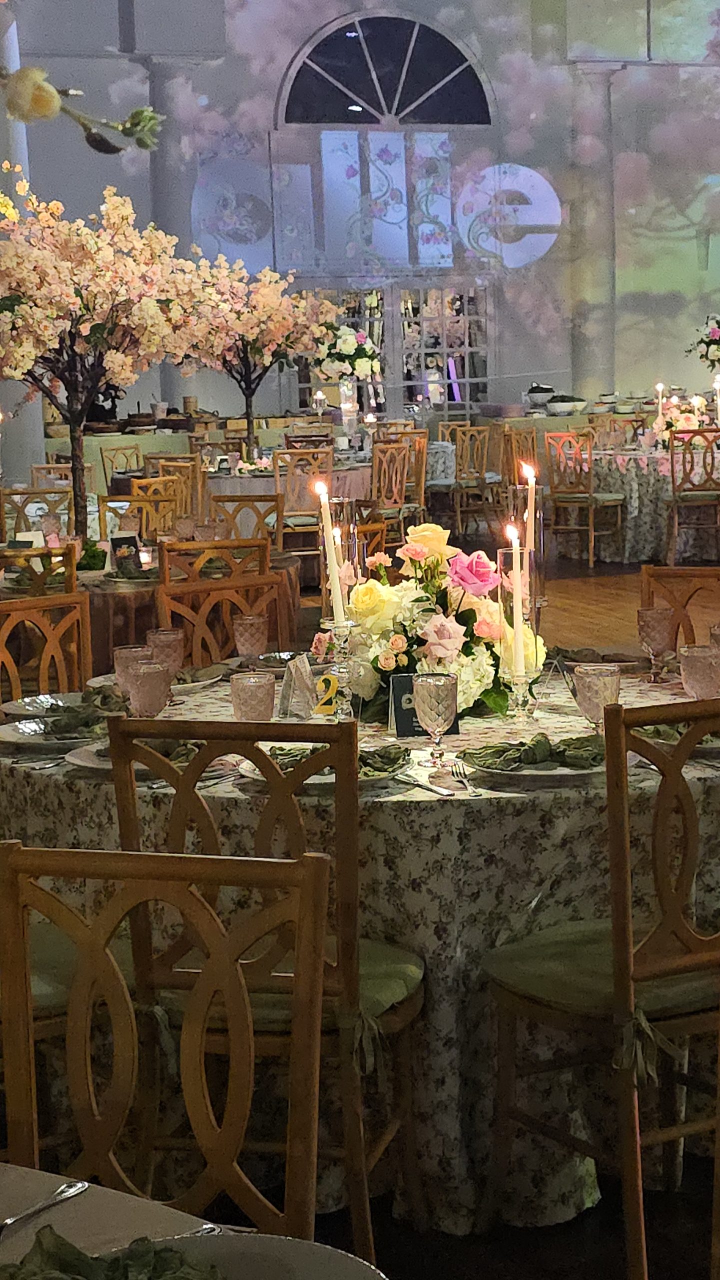 A blossom tree over the set tables, the projected garden and gate on the wall behind, the girl's name above it