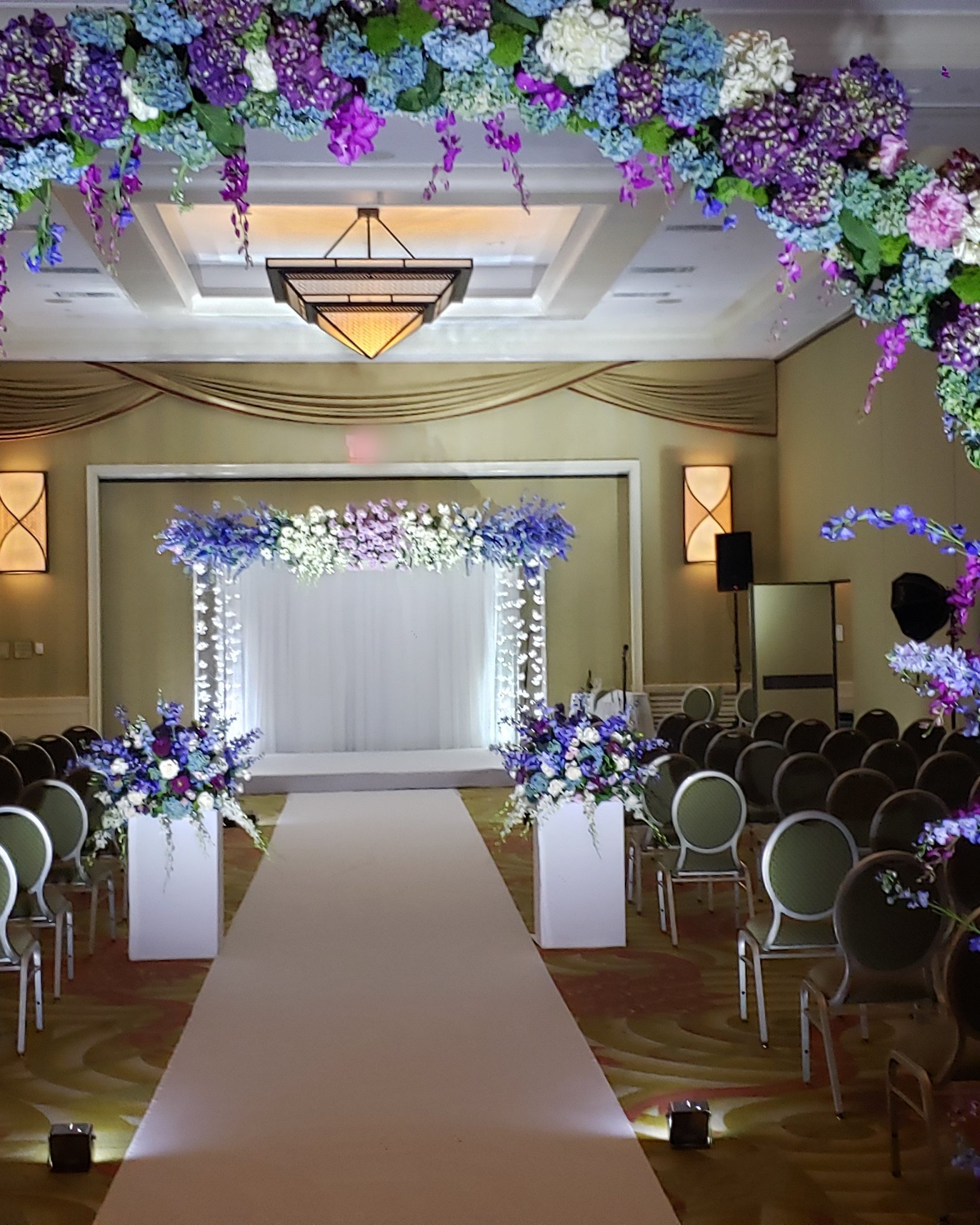 Looking through a lavender and blue floral arch down a white aisle to a chuppah lit white
