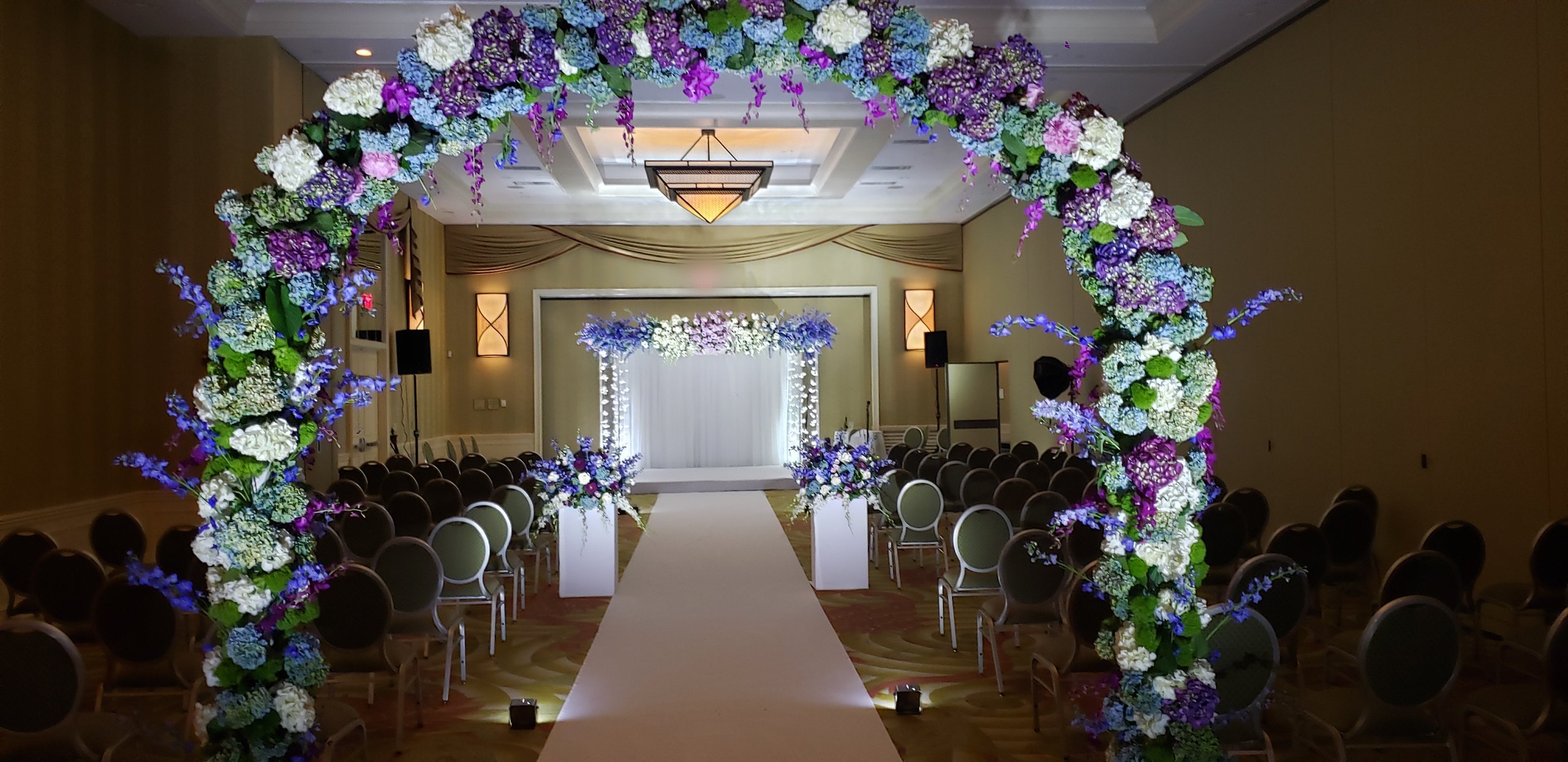 The whole ceremony room: the floral arch, the chairs, the white aisle and the chuppah at the end