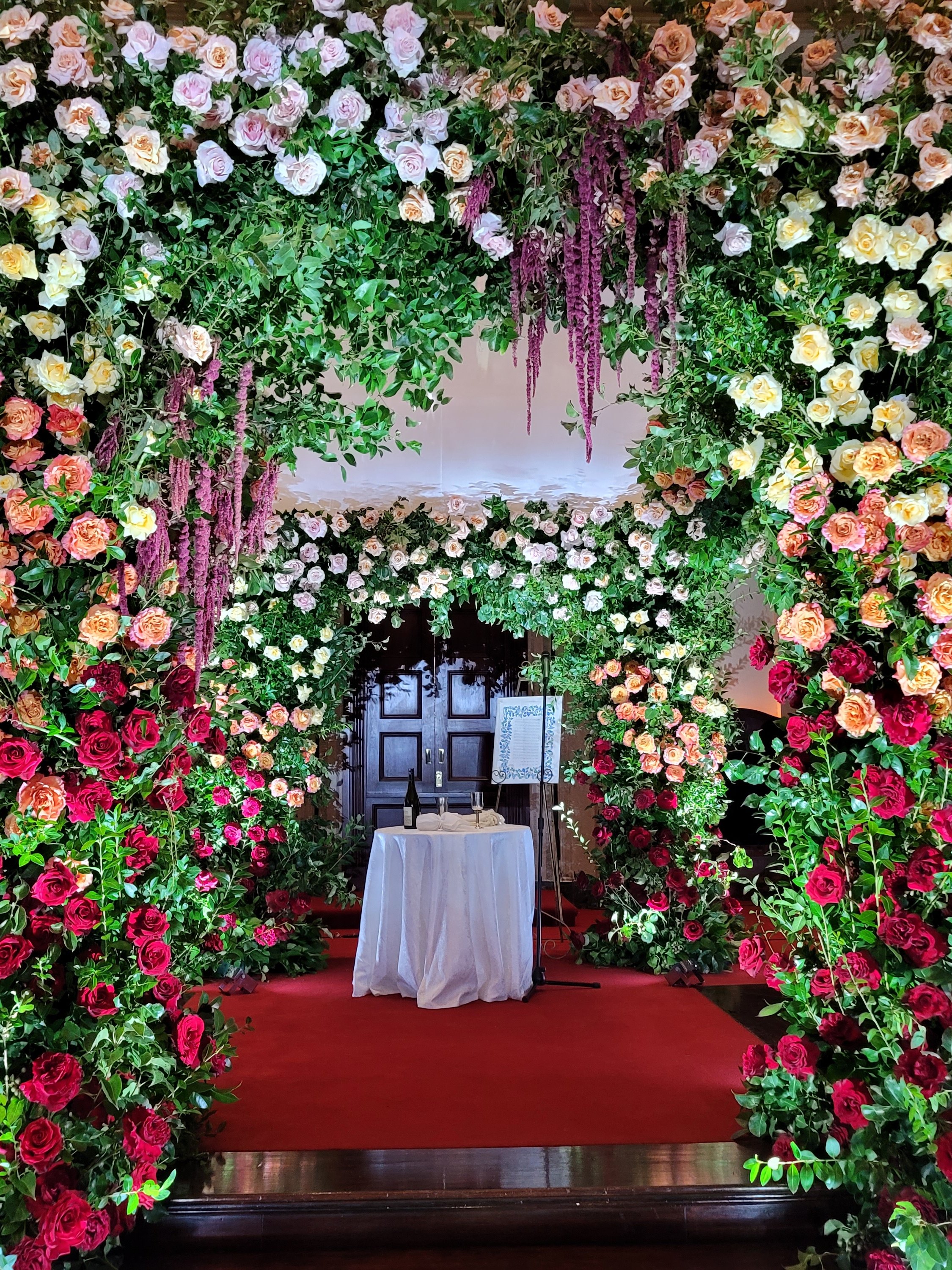 A chuppah built of roses and greenery in pink, peach and red, a red carpet leading to a small white table