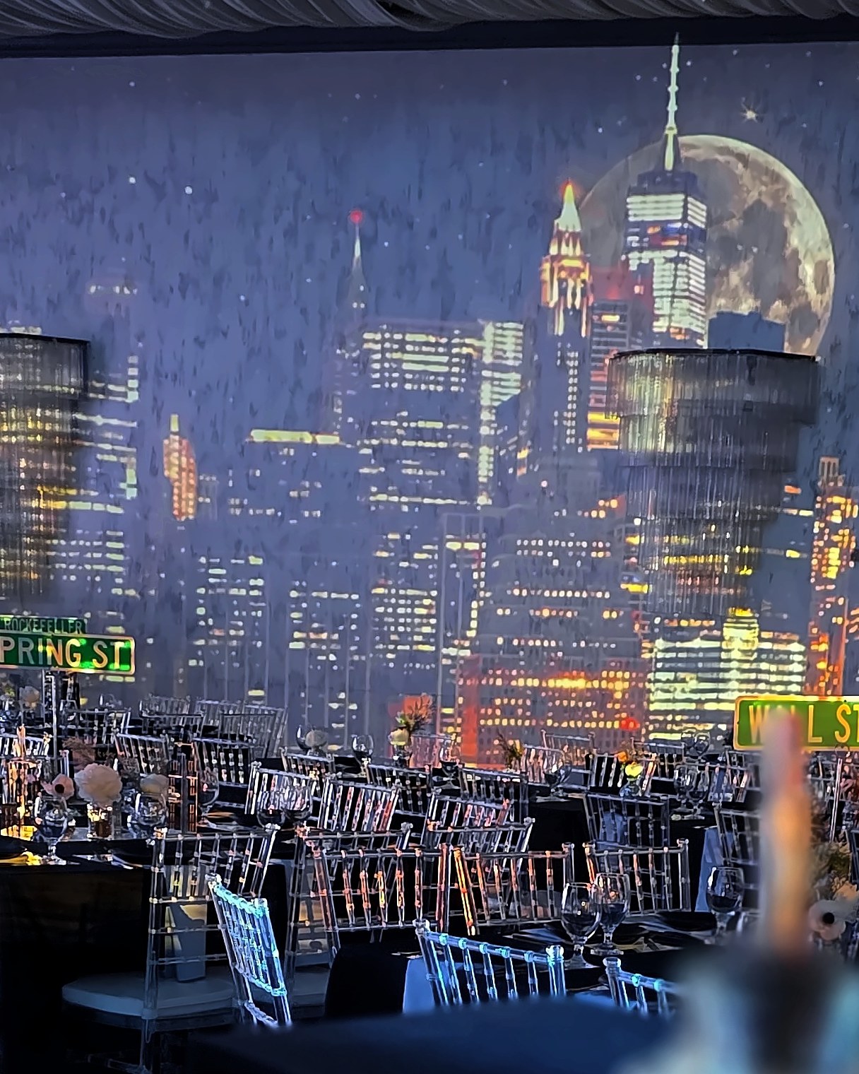The whole back wall lit as the New York skyline at night, a full moon, crystal chandeliers and street signs over the set tables