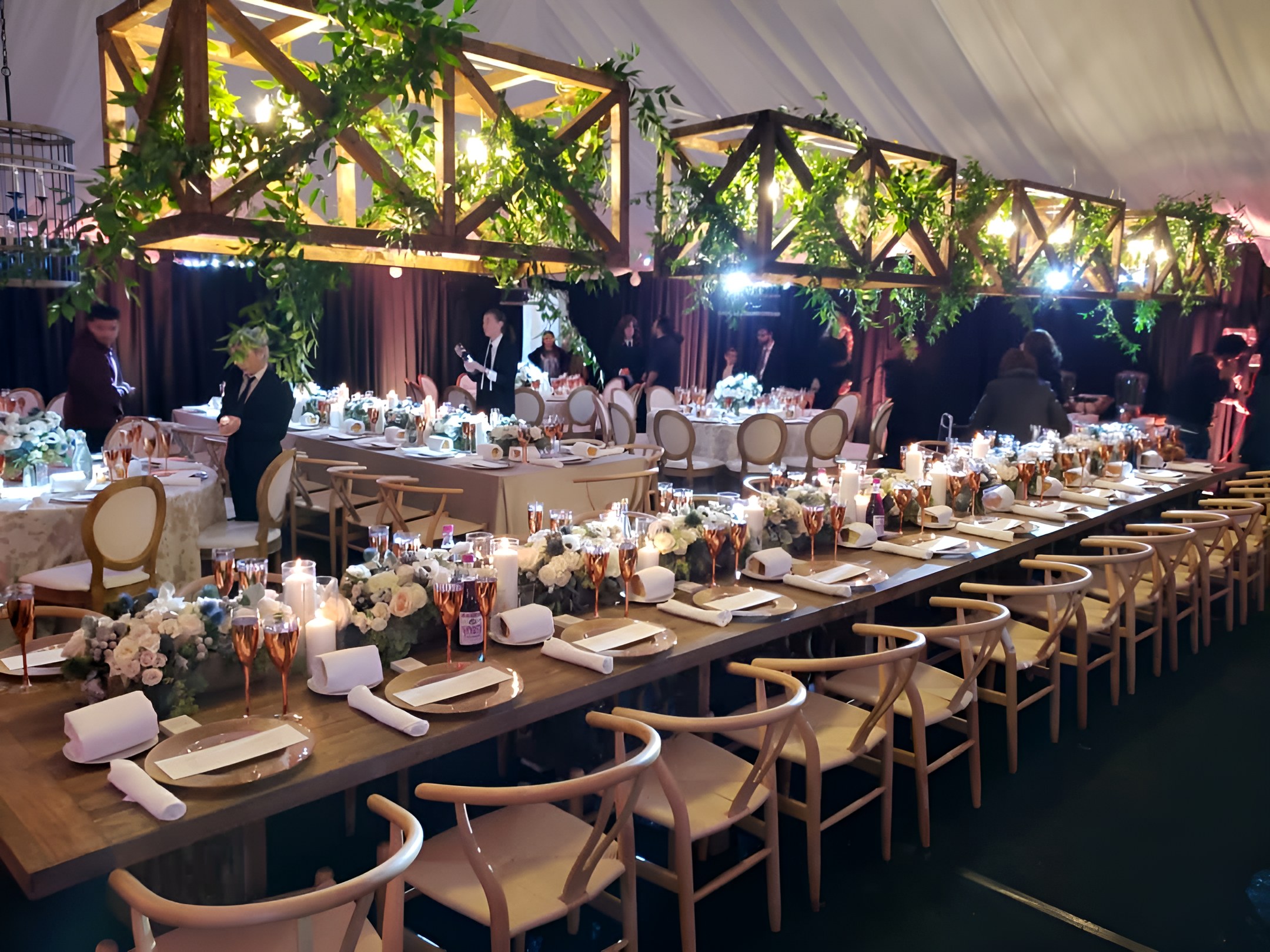 A warm tent for a Friday-night dinner: timber-and-greenery beams lit over the long tables