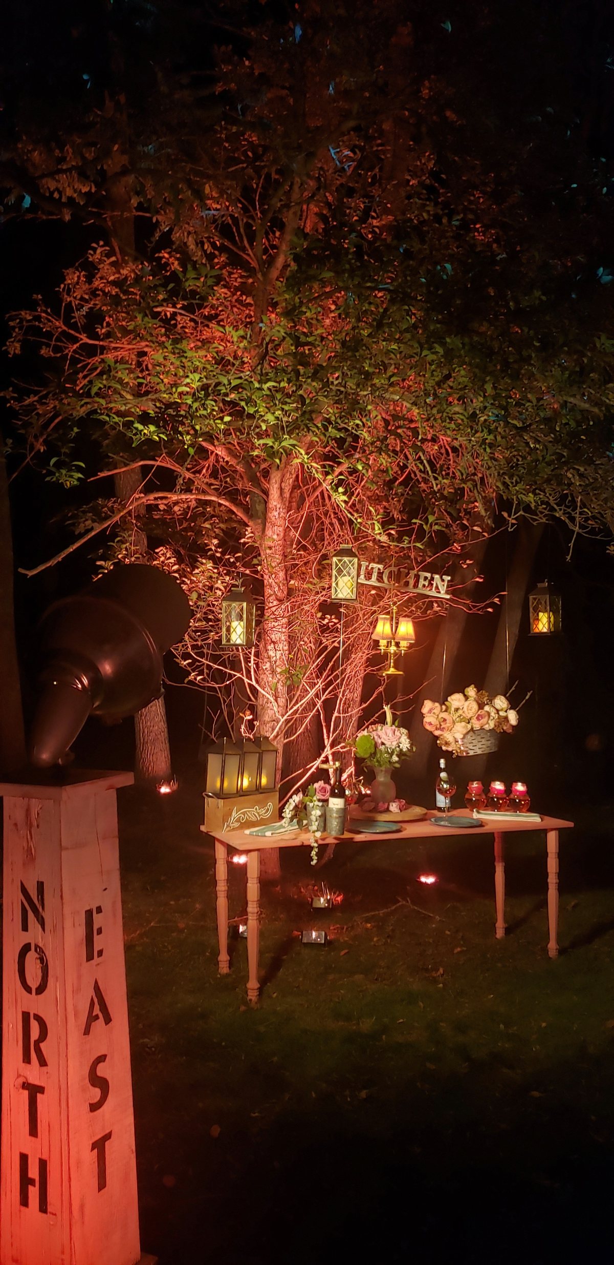 A tree lit red at night over a lantern-lit table on the lawn